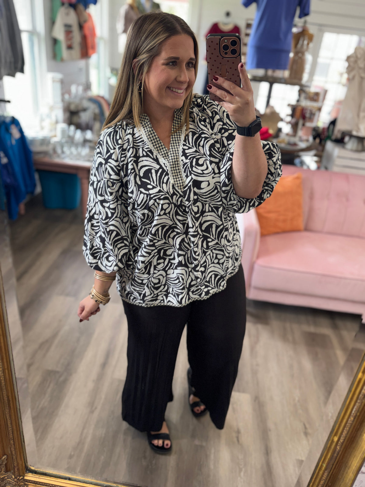 A woman takes a mirror selfie in a clothing boutique, wearing a black and white abstract patterned blouse with three-quarter sleeves and black wide-leg pants.