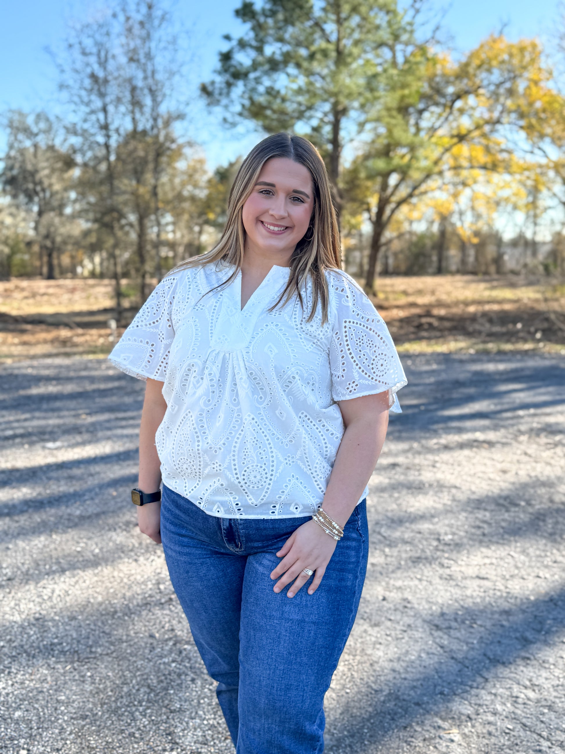 Woman wearing a white blouse and blue jeans standing on a gravel road with trees in the background.
