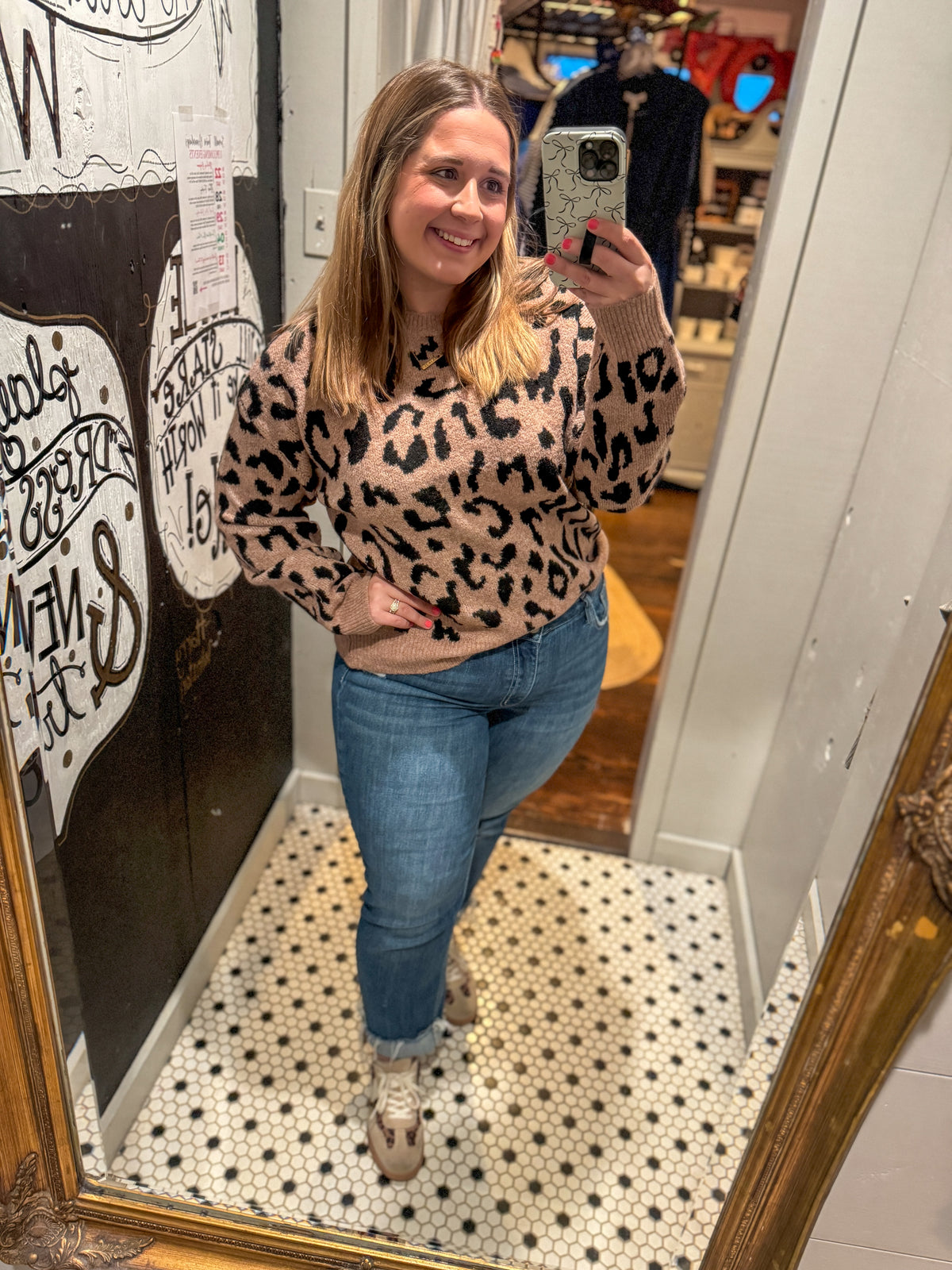 Woman taking a mirror selfie wearing a leopard print sweater and blue jeans in a store.