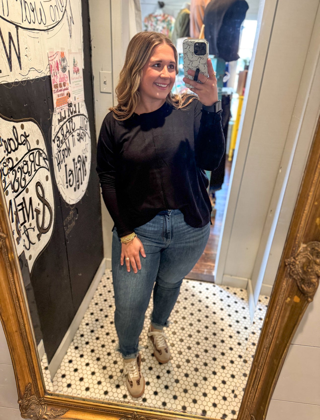 Woman taking a mirror selfie wearing a black sweater and blue jeans in a store.