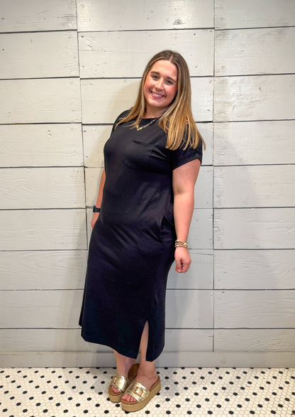 Woman wearing a black dress standing against a light wooden paneled wall.