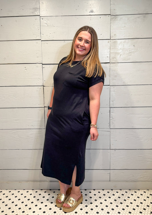 Woman wearing a black dress standing against a light wooden paneled wall.