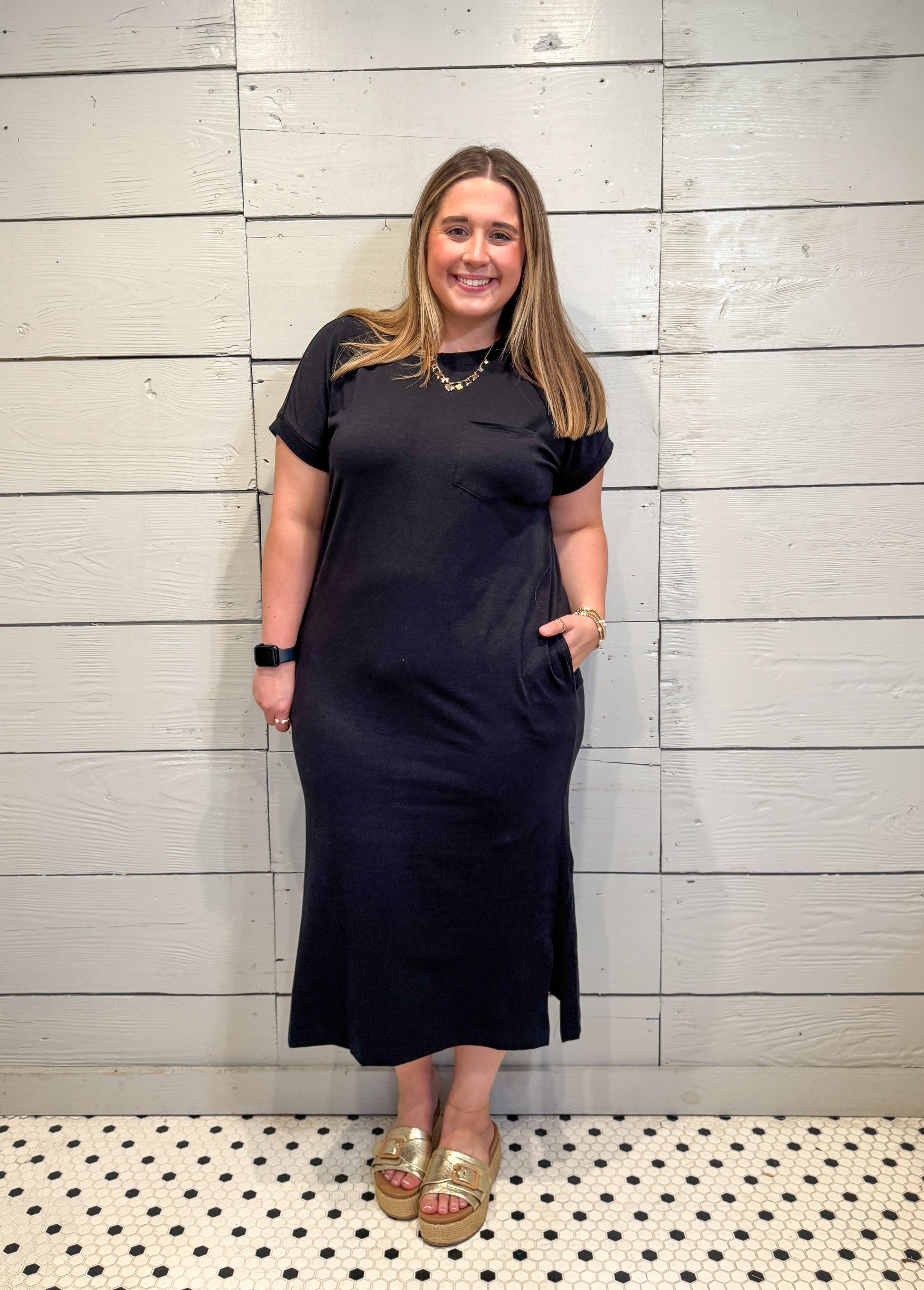 Woman wearing a black dress standing against a light wooden paneled wall.