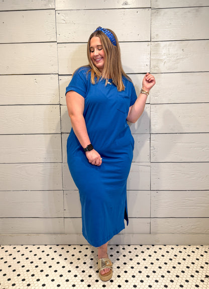 Woman wearing a blue dress standing against a light wooden paneled wall.