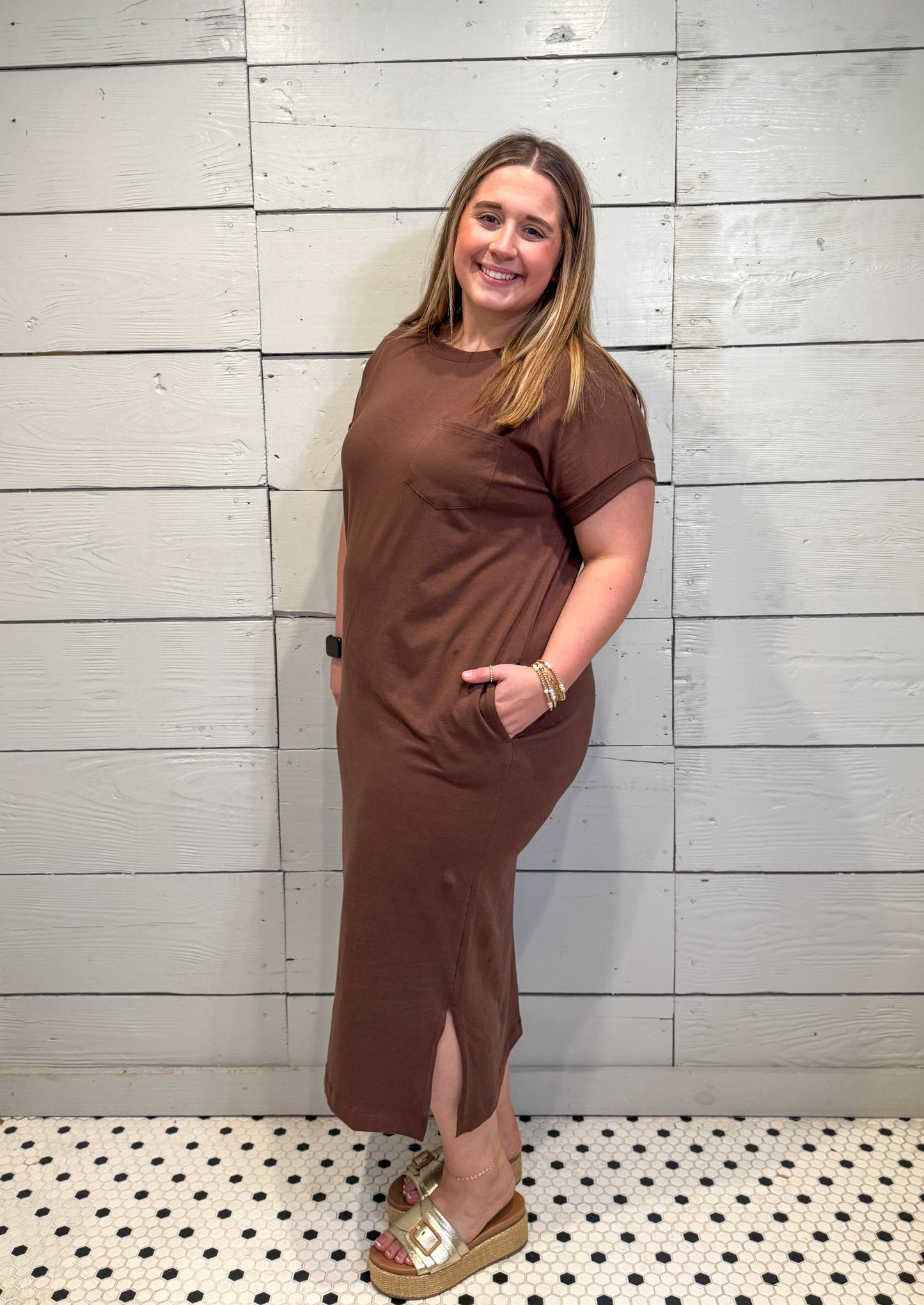 Woman wearing a brown dress standing against a light wooden panel wall.