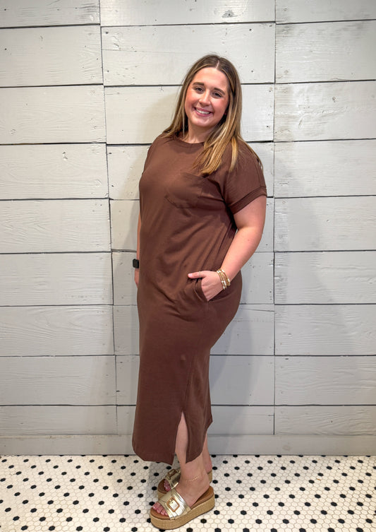 Woman wearing a brown dress standing against a light wooden panel wall.