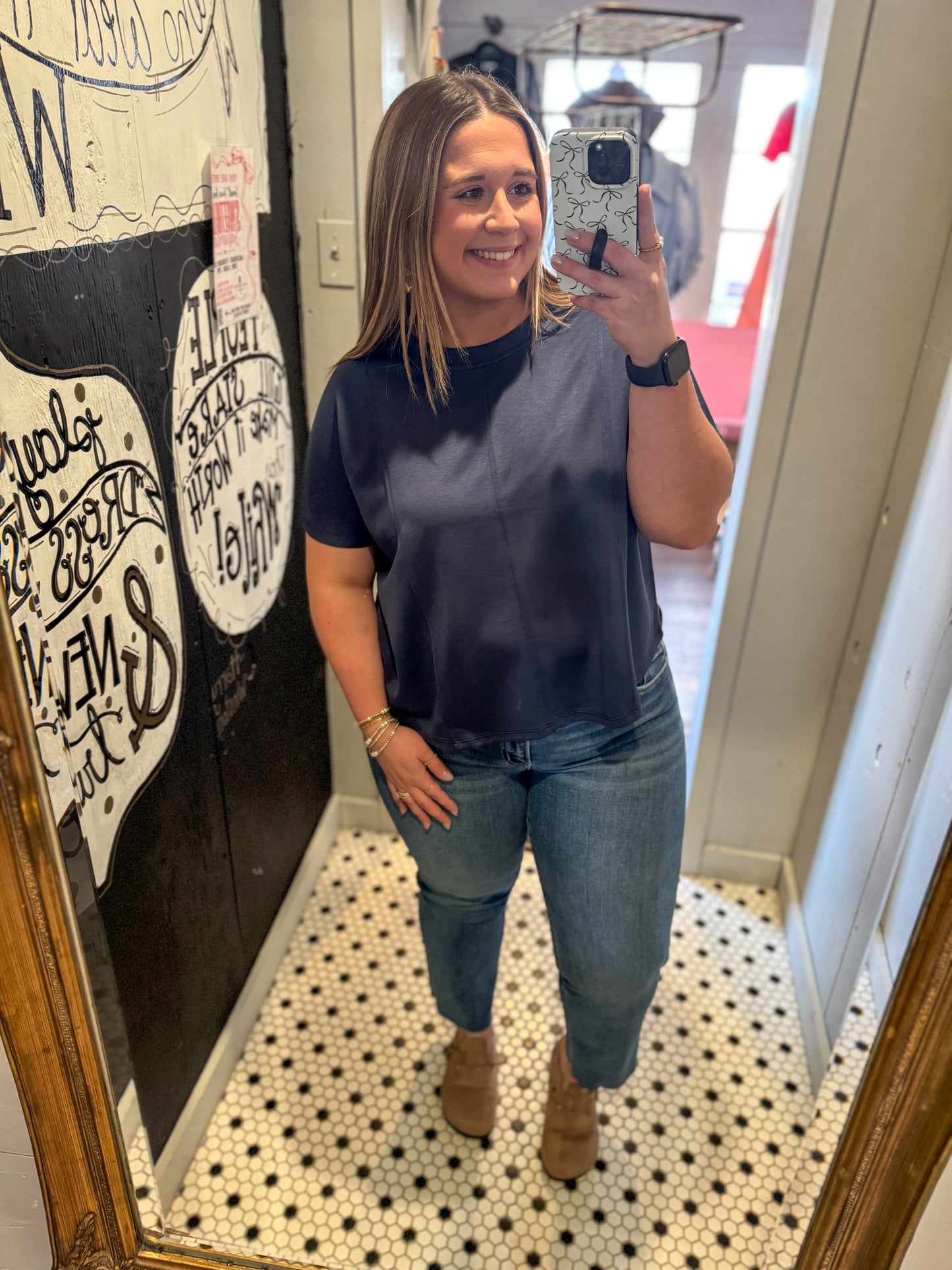 Woman taking a mirror selfie wearing a navy blue top and jeans in a room with decorative elements.