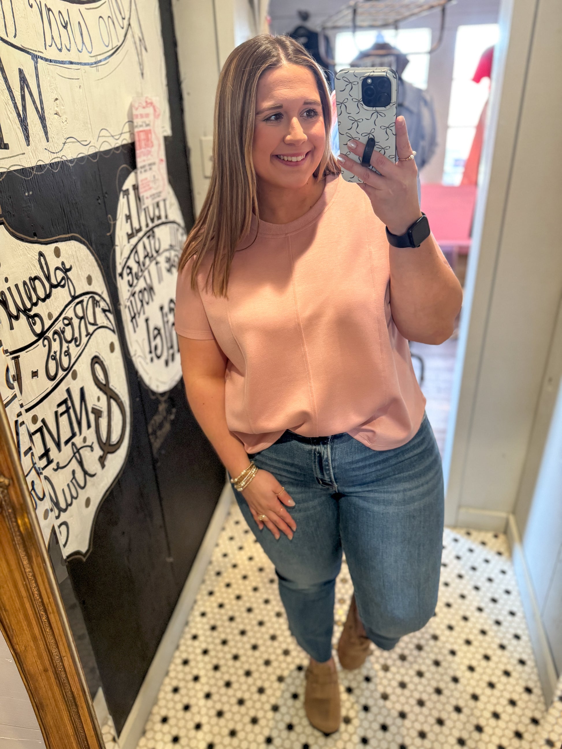 Woman taking a mirror selfie wearing a pink top and jeans in a store.