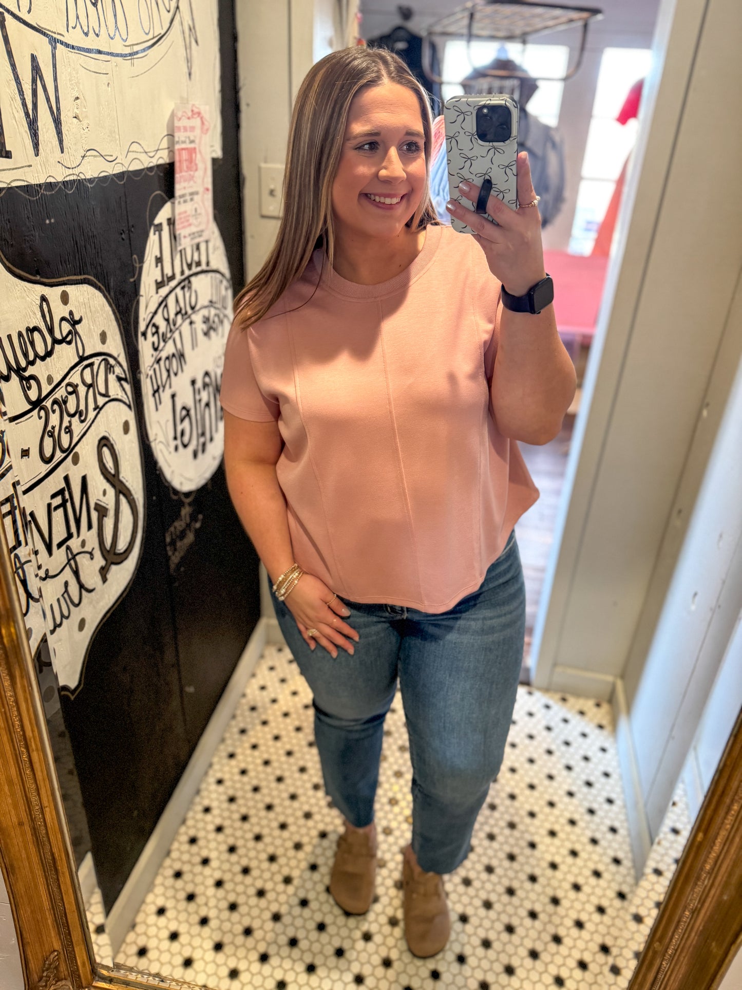 Woman taking a mirror selfie wearing a pink top and jeans indoors.
