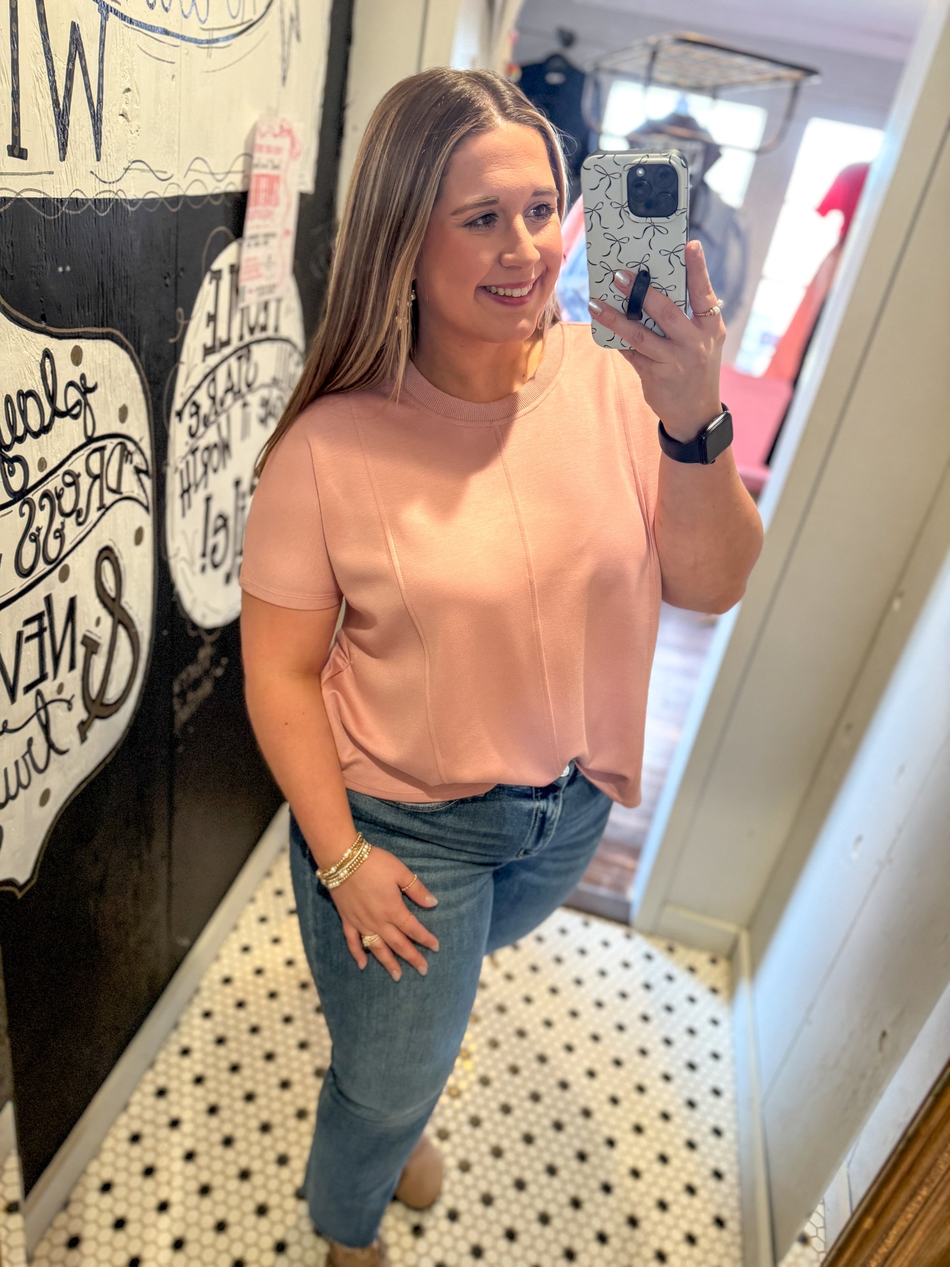 Woman taking a mirror selfie wearing a pink top and blue jeans in a casual setting.