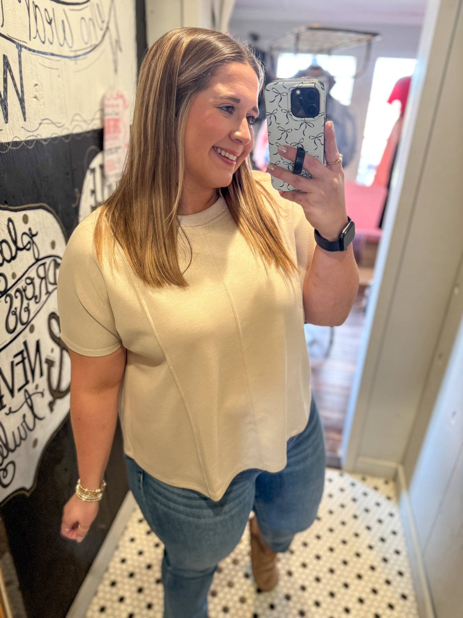 Woman taking a mirror selfie wearing a beige top and blue jeans in a room with a decorative wall.