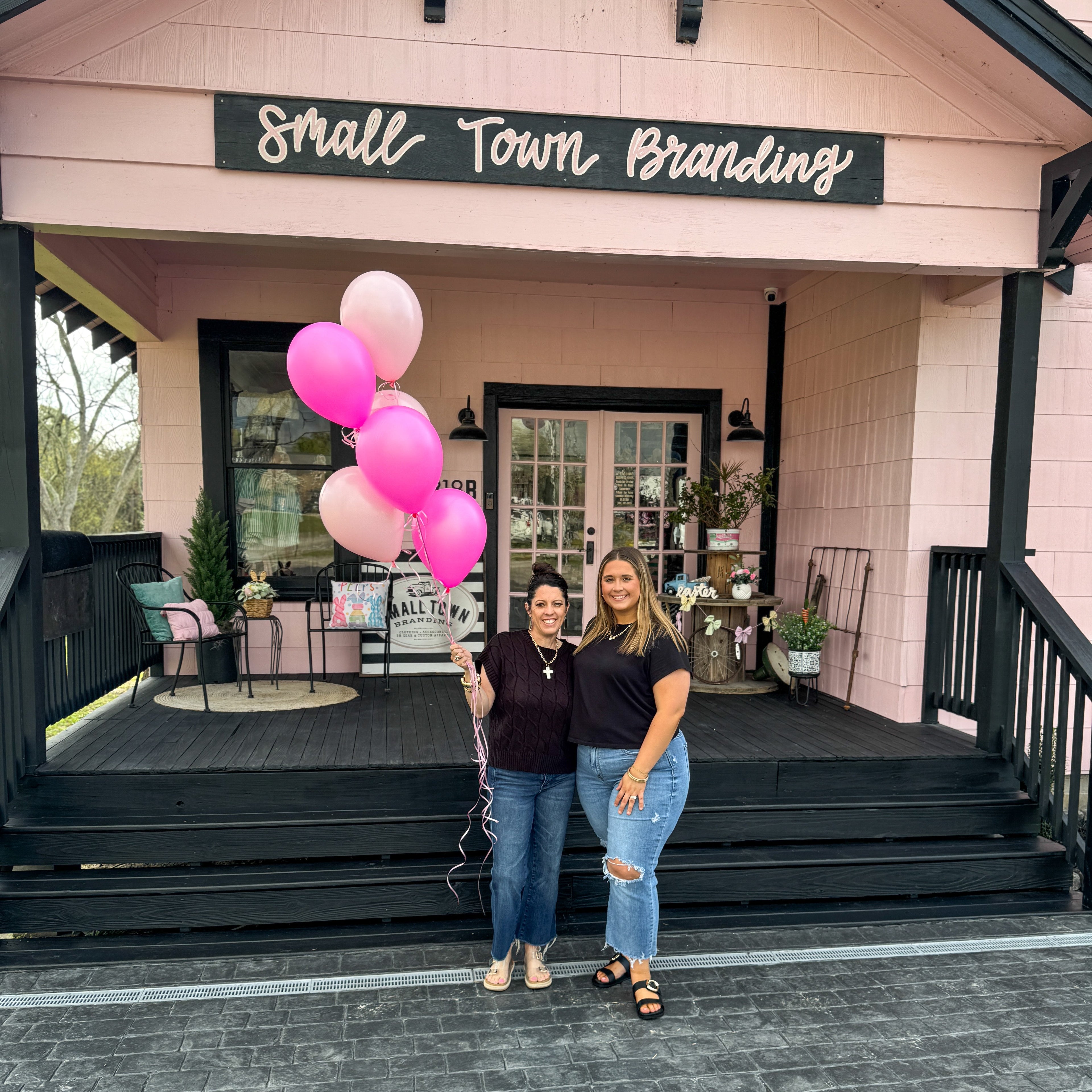 Two women standing in front of a building with 'Small Town Branding' sign, holding pink balloons.