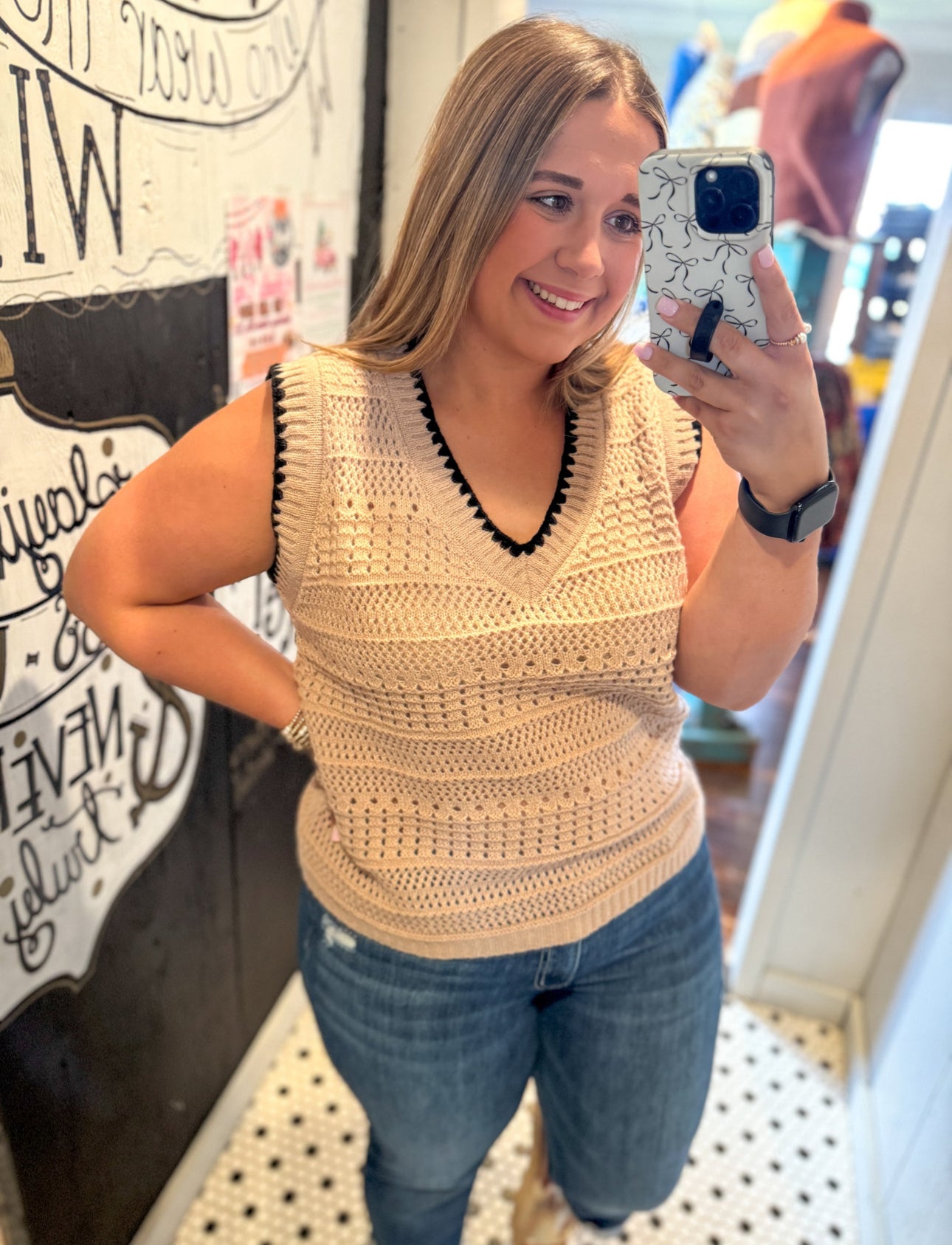 Woman taking a mirror selfie wearing a beige crochet top and blue jeans.
