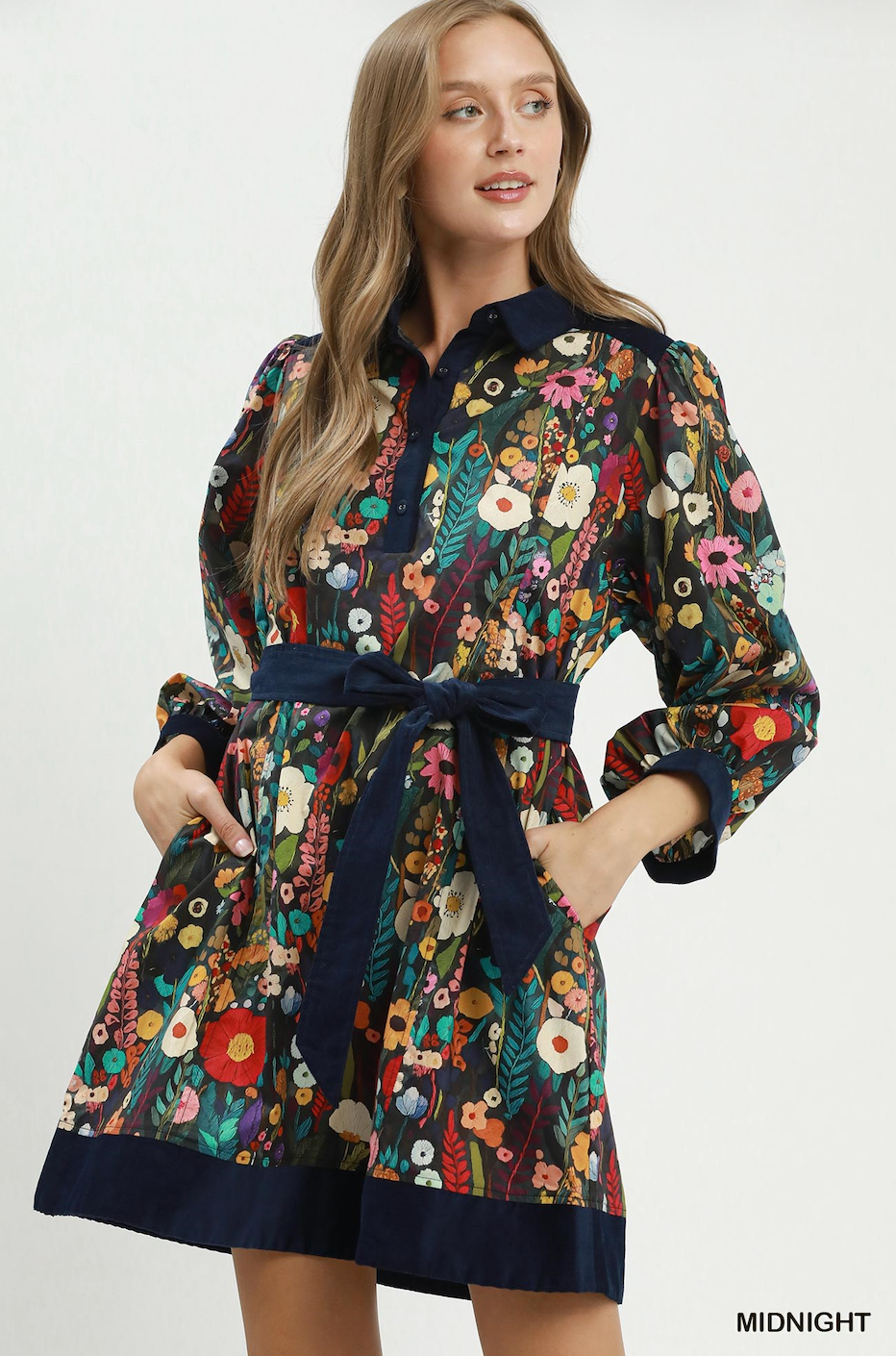 A woman modeling a short floral print shirt dress with a navy blue collar, waist tie belt, and hem. The dress features a multicolor floral pattern on a dark background with navy blue trim.