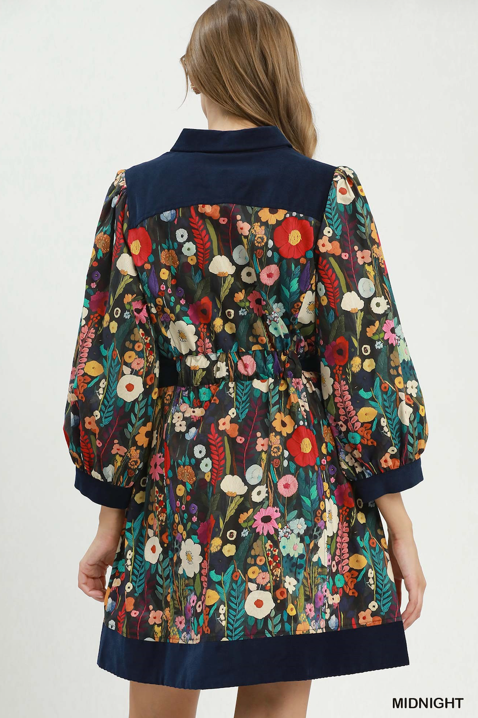 A woman shown from the back wearing a floral print dress with dark navy blue trim on the collar, cuffs, and hem. The floral pattern features red, yellow, pink, and white flowers on a dark background.