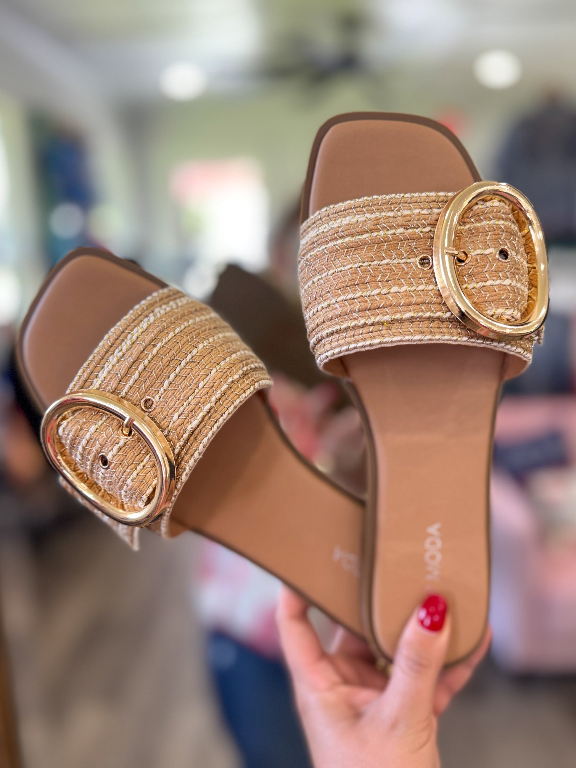 Pair of brown sandals with gold buckles held by a hand against a blurred indoor background