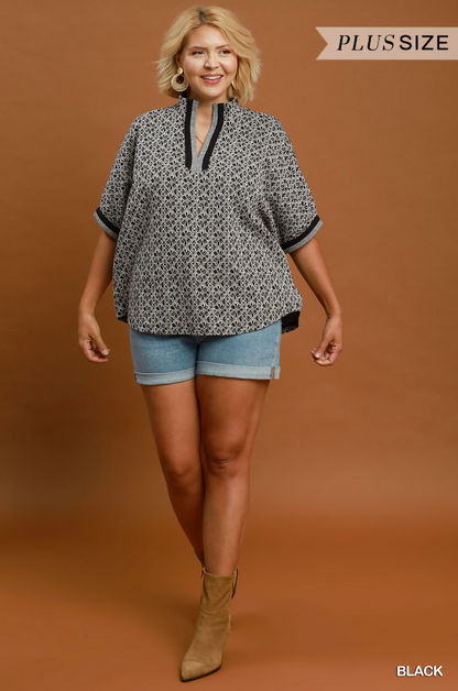 A woman modeling a black and white geometric patterned short-sleeve blouse with black trim, paired with denim shorts and tan ankle boots, against a solid brown studio background. A 'PLUS SIZE' banner is visible in the top right, and 'BLACK' is written in the bottom right.