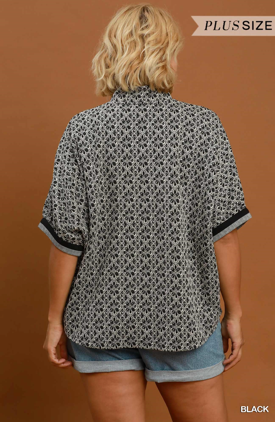 A woman shown from the back wearing a loose short-sleeve button-up shirt with a black and white geometric pattern and rolled cuffs, paired with denim shorts, against a warm brown studio background. Text in the top right reads 'PLUSSIZE' and 'BLACK' appears in the bottom right corner.