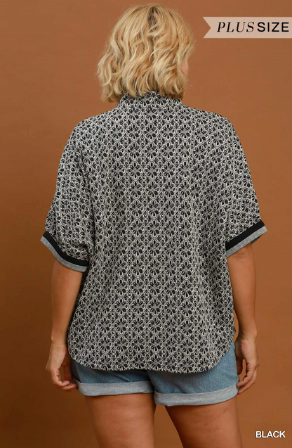 A woman shown from the back wearing a loose short-sleeve button-up shirt with a black and white geometric pattern and rolled cuffs, paired with denim shorts, against a warm brown studio background. Text in the top right reads 'PLUSSIZE' and 'BLACK' appears in the bottom right corner.