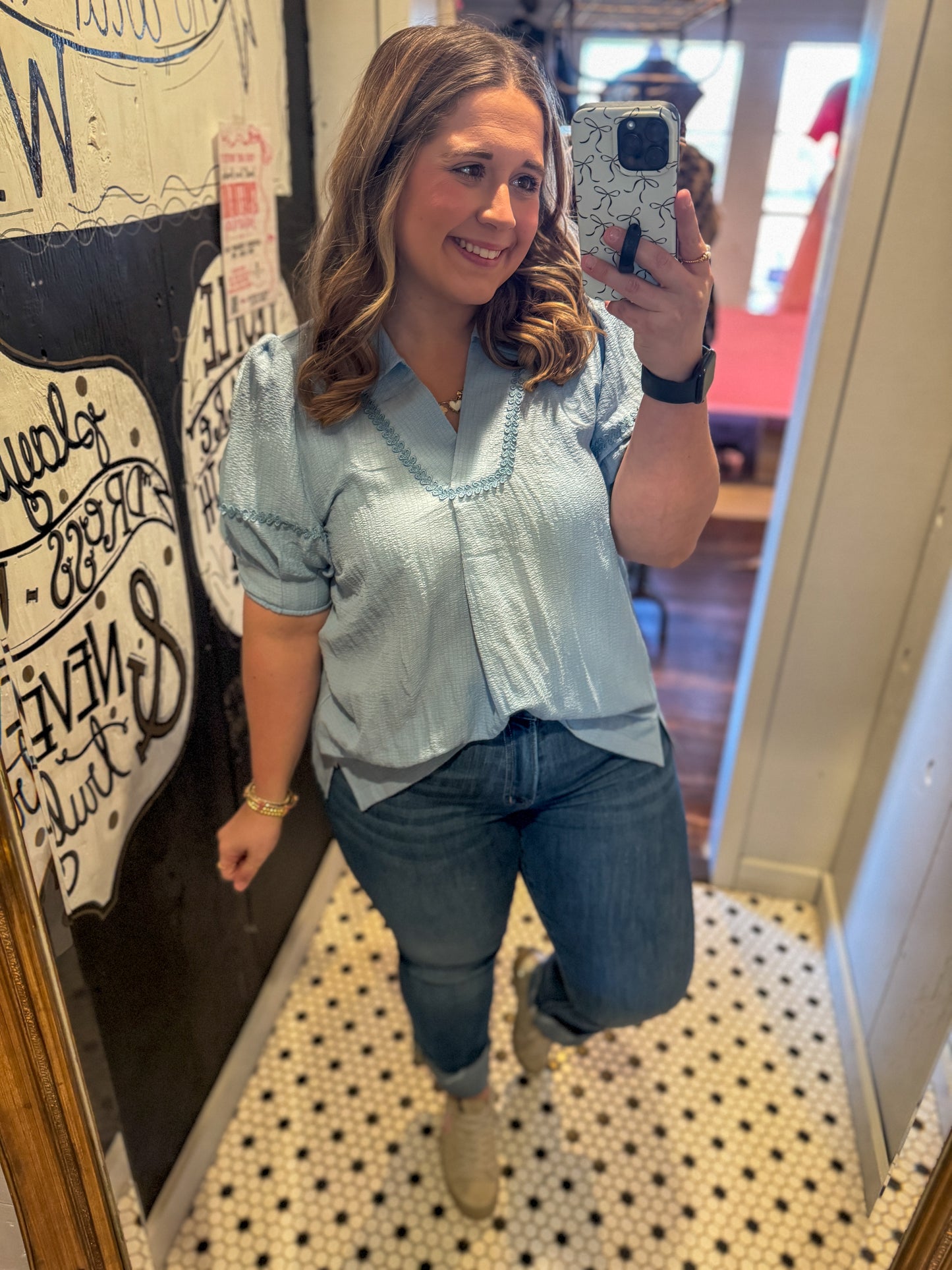 Woman taking a mirror selfie wearing a light blue blouse and jeans.