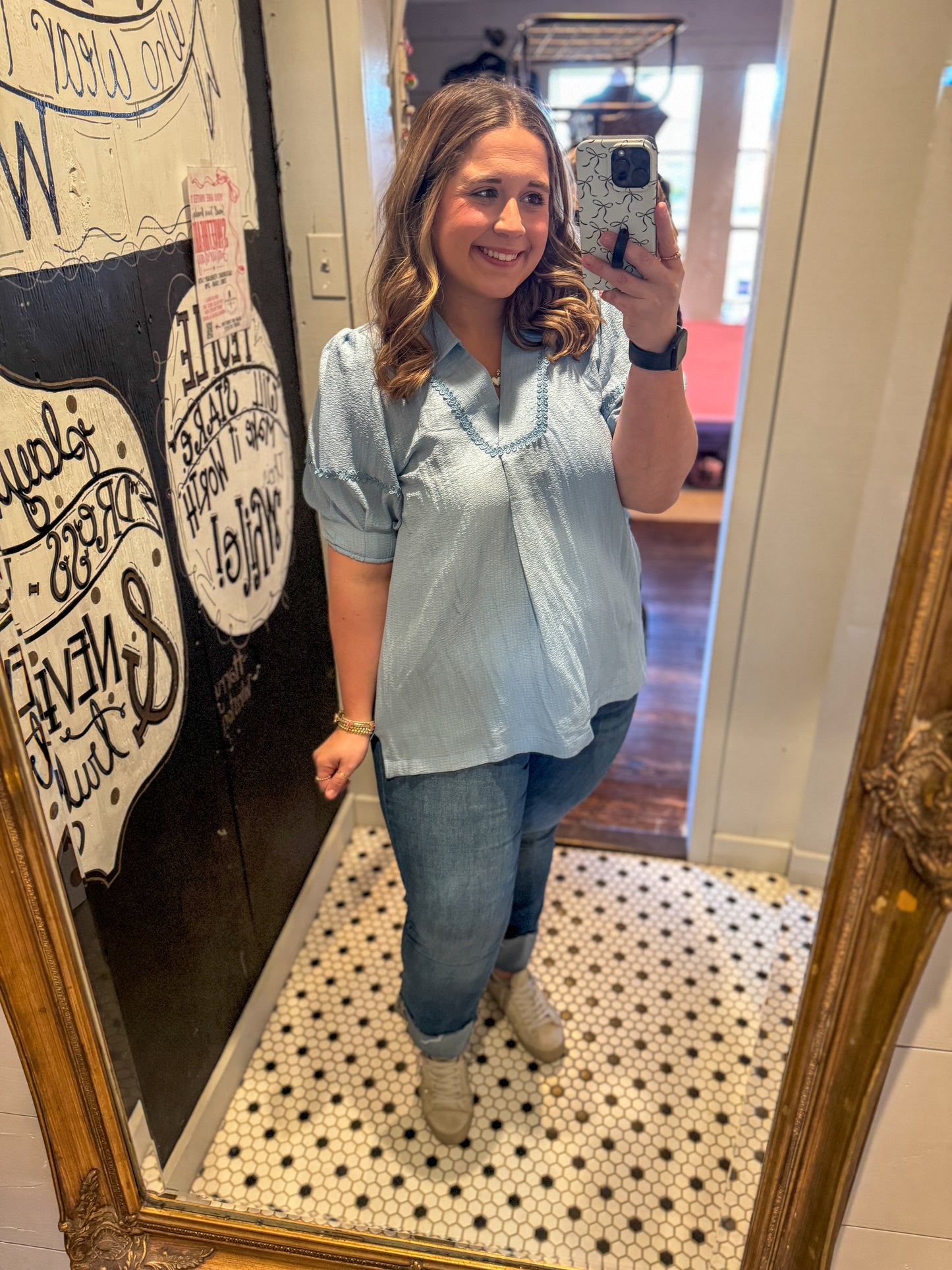 Woman taking a mirror selfie wearing a light blue shirt and jeans in a room with decorative elements.