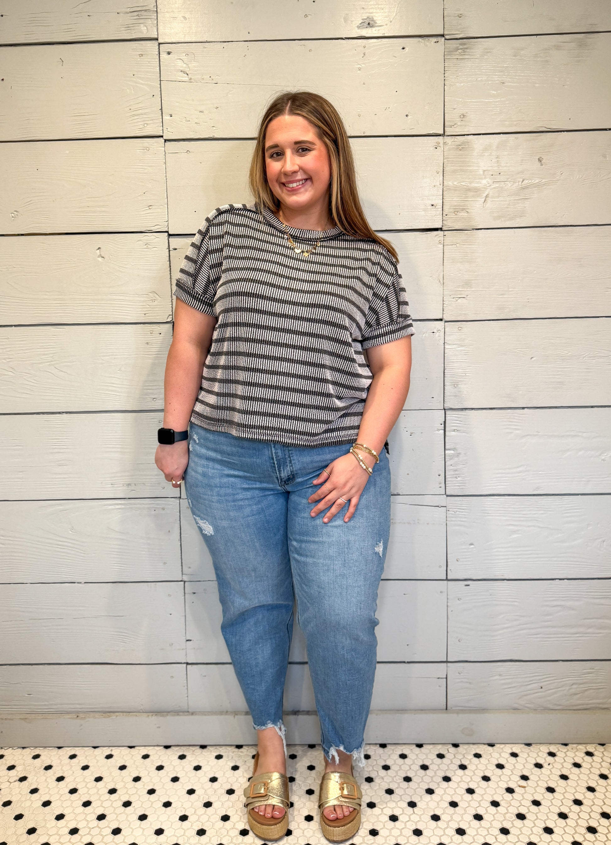 Woman wearing a striped shirt and jeans standing against a wooden wall.