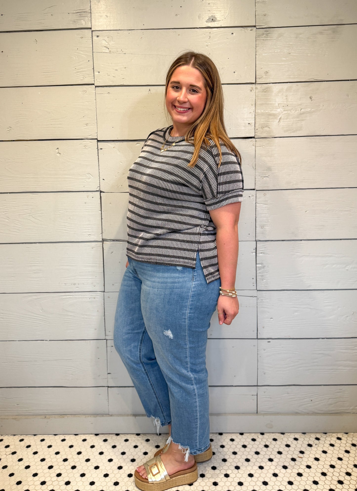 Woman wearing a striped shirt and jeans standing against a wooden paneled wall.