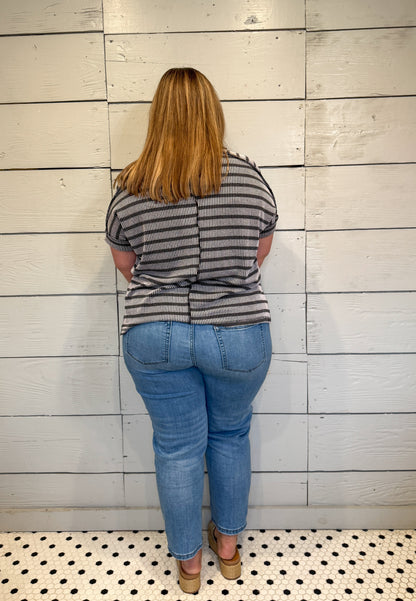 Person wearing a striped shirt and blue jeans standing against a light wooden paneled wall.