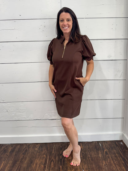 A woman modeling a brown short-sleeve shift dress with puff sleeves and a gold zipper at the collar, standing against a white shiplap wall and dark wood floor.