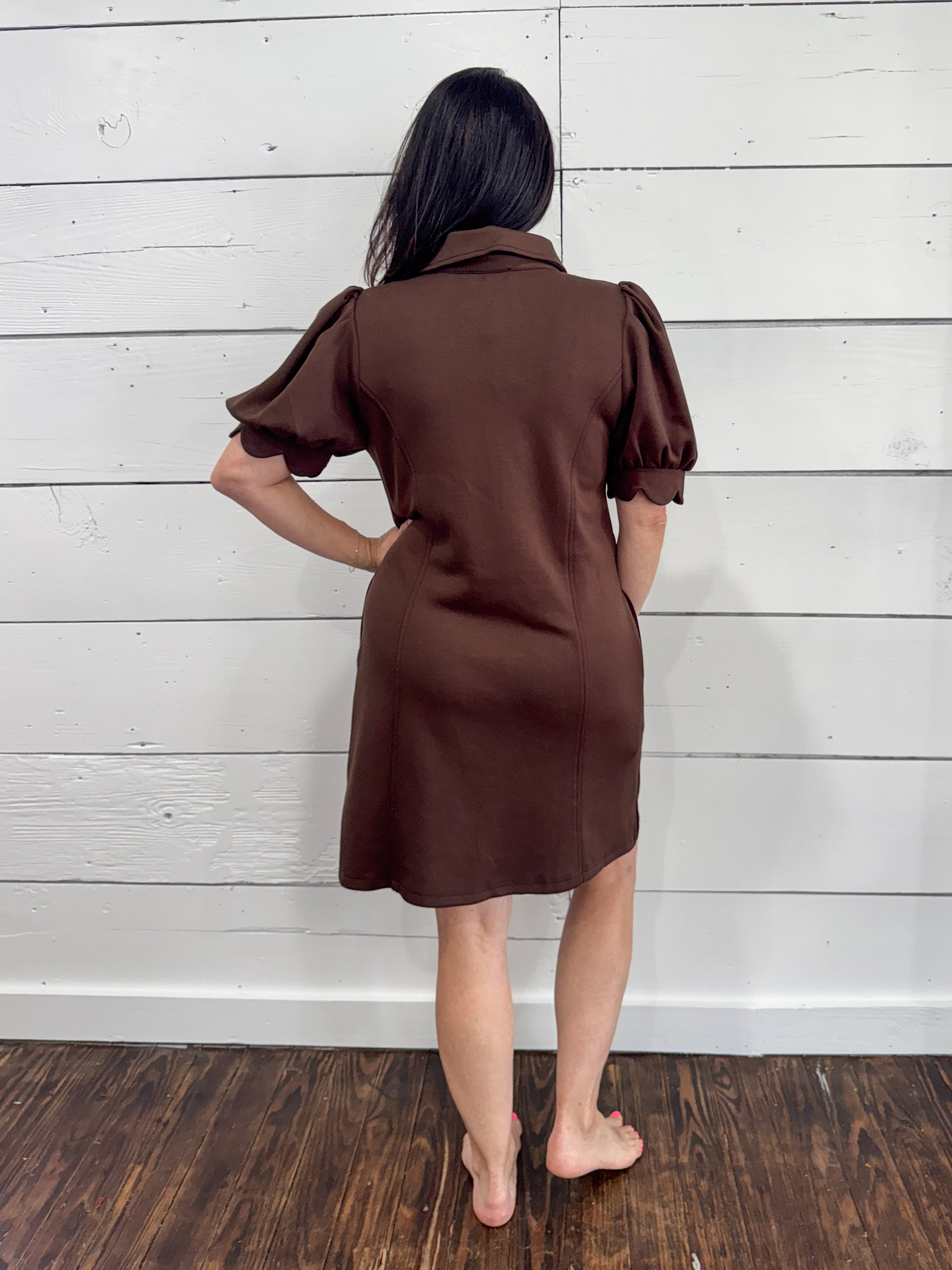 A woman stands with her back to the camera wearing a brown short-sleeve collared dress with puffed sleeves, set against a white wood-paneled wall and dark wood floor.