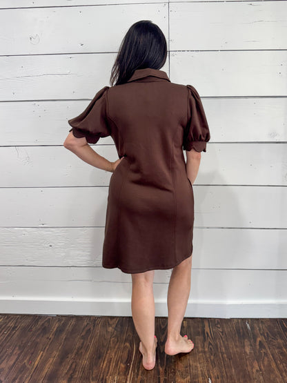 A woman stands with her back to the camera wearing a brown short-sleeve collared dress with puffed sleeves, set against a white wood-paneled wall and dark wood floor.