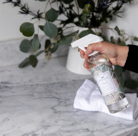 A hand holding a clear spray bottle labeled 'FRESH AIR' and spraying a mist onto a white cloth on a marble countertop, with green foliage in the background.