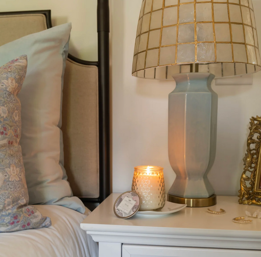A white scented candle in a textured clear glass jar with a patterned metal lid leaning against it, photographed on a white side table with a lap beside it.