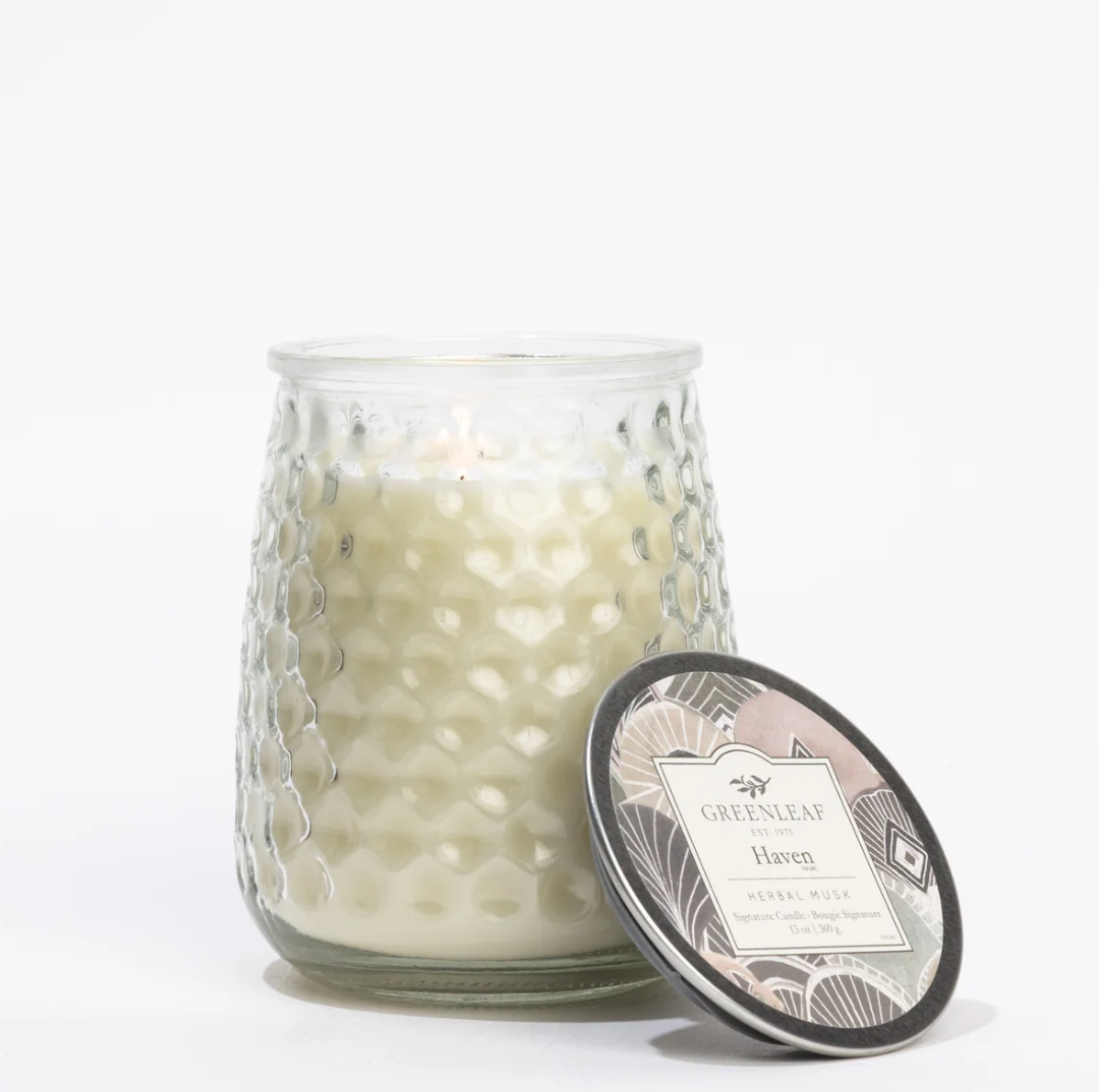 A white scented candle in a textured clear glass jar with a patterned metal lid leaning against it, photographed on a clean white background. The label reads Greenleaf Haven.
