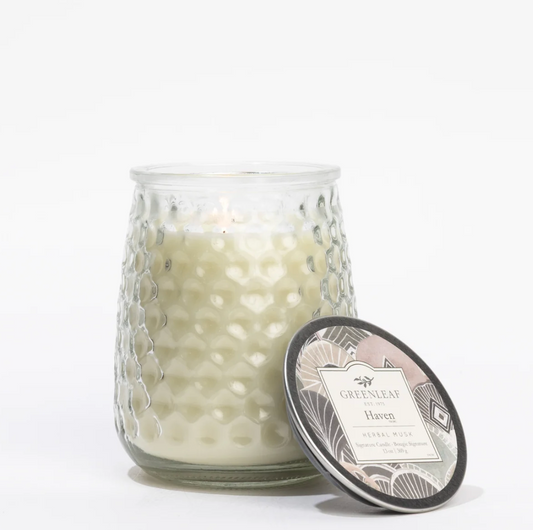 A white scented candle in a textured clear glass jar with a patterned metal lid leaning against it, photographed on a clean white background. The label reads Greenleaf Haven.