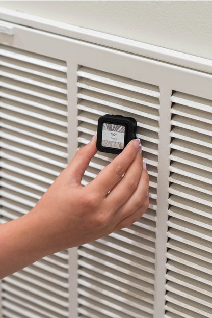 Hand holding a small black device with a screen in front of a white radiator.