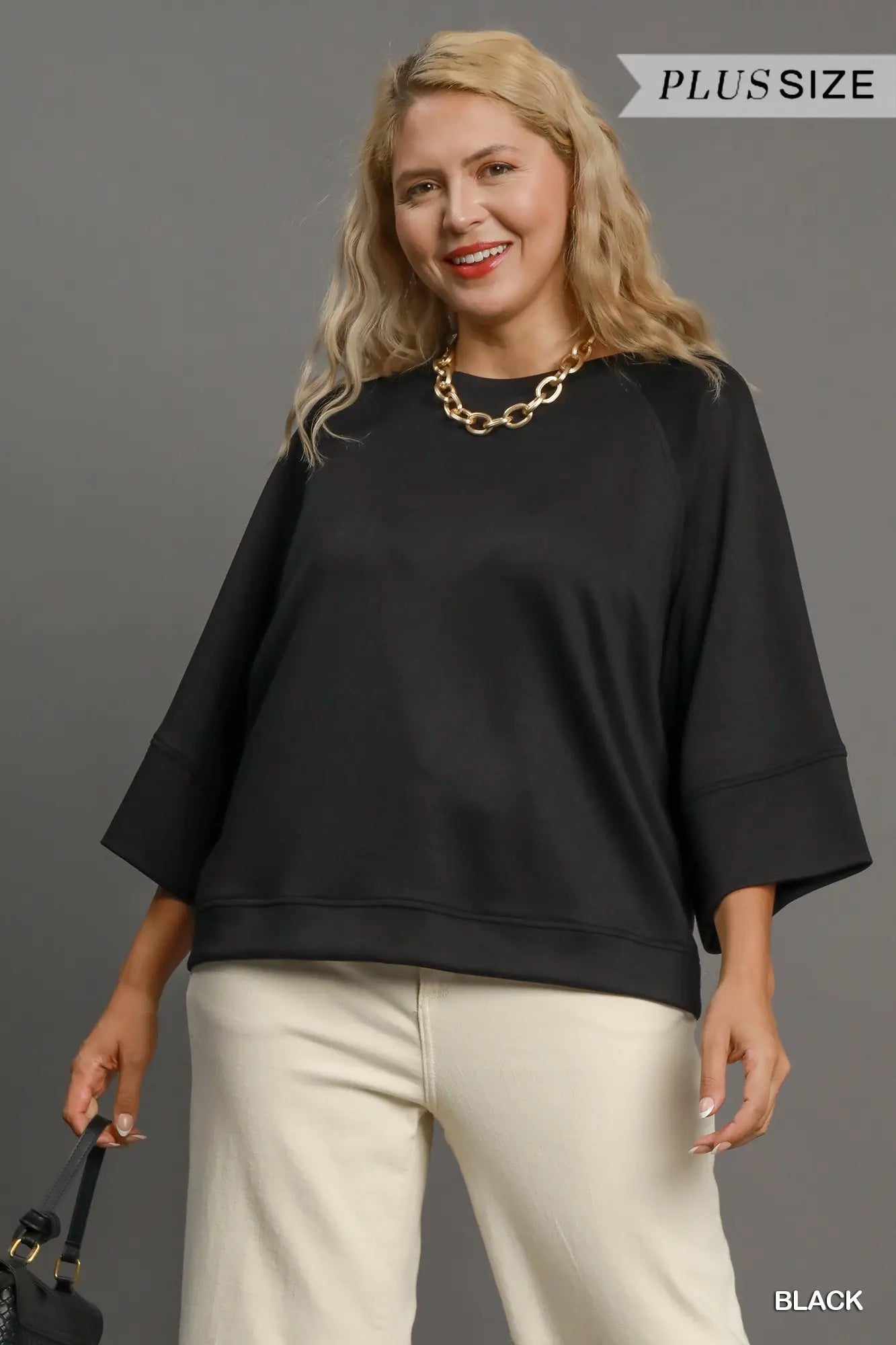 Woman wearing a black plus-size top and white pants against a gray background