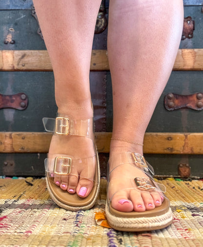 Transparent sandals with gold buckles worn by a person on a textured rug with vintage suitcases in the background.