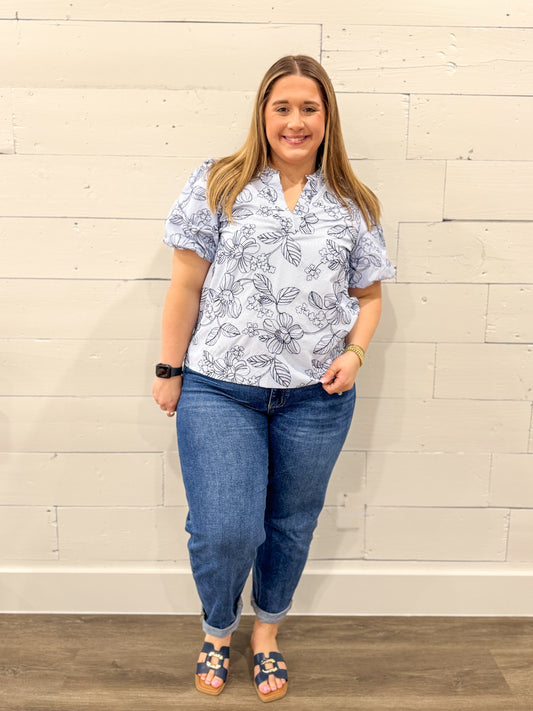 Woman wearing a floral blouse and jeans standing against a white brick wall.