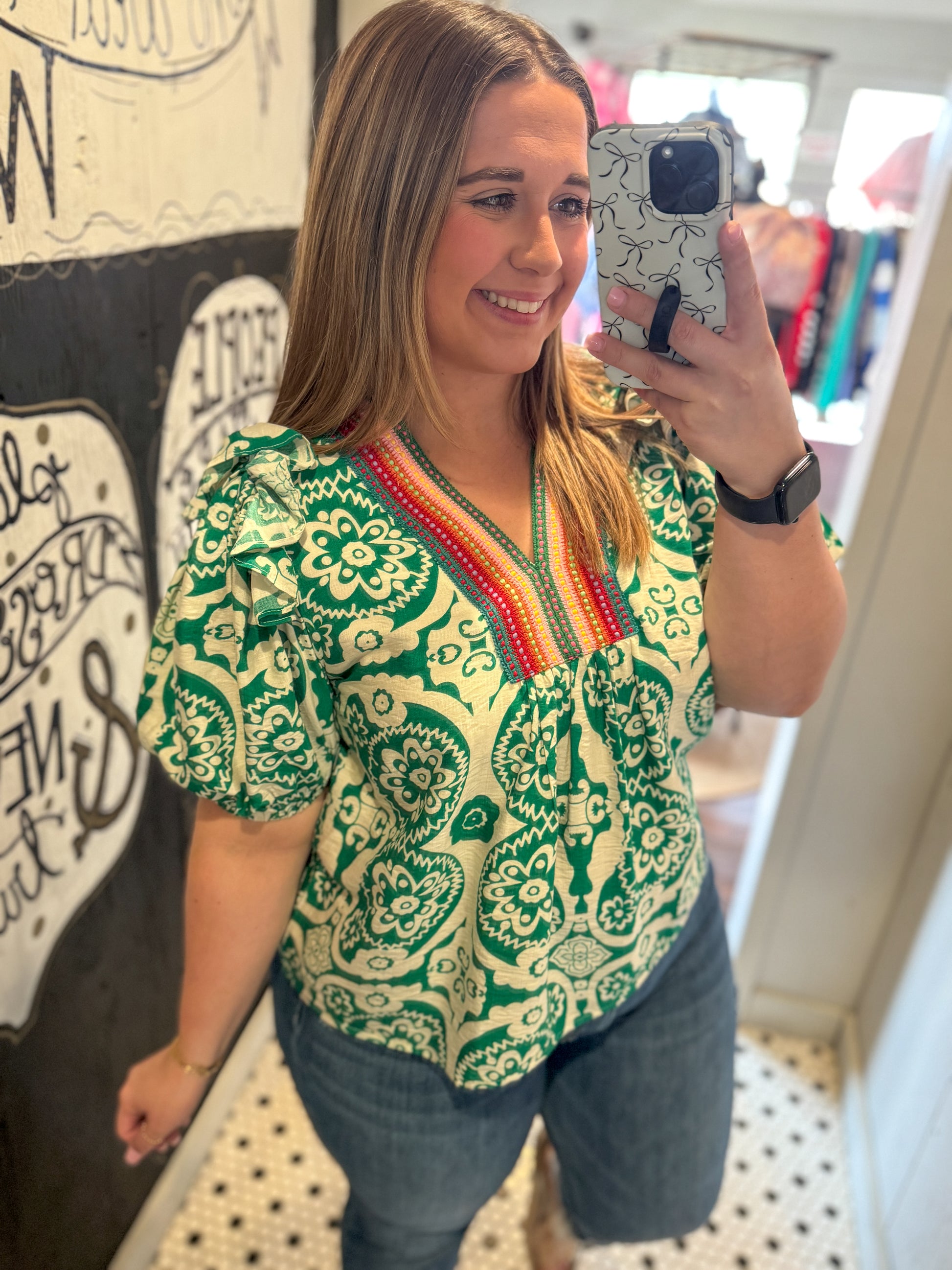 Woman taking a mirror selfie wearing a green and white patterned top with colorful trim.