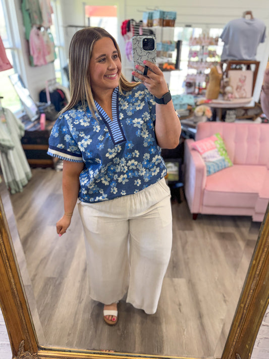 Woman taking a mirror selfie wearing a blue floral blouse and white pants in a store.