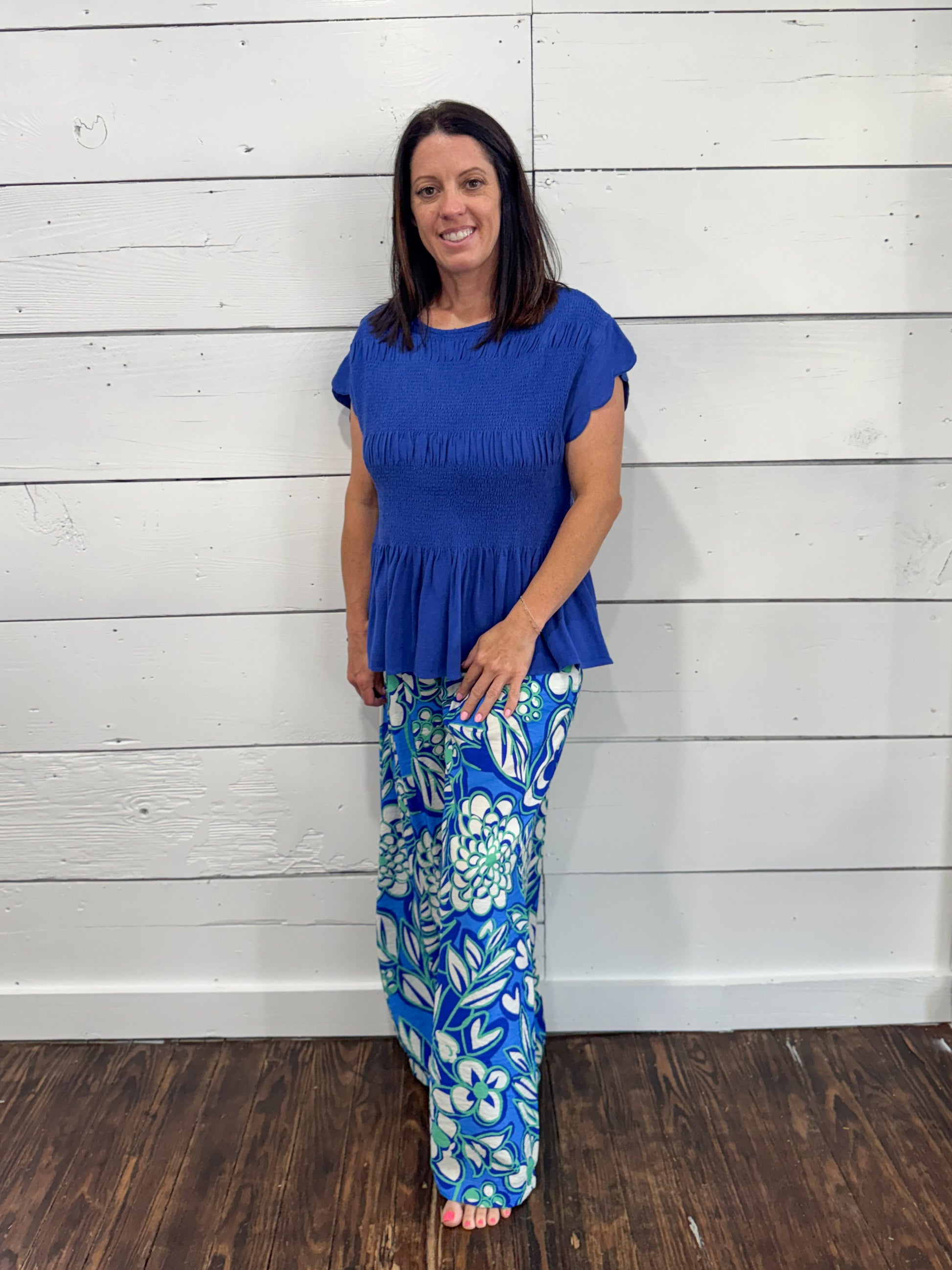 A woman modeling a two-piece outfit consisting of a royal blue short-sleeve peplum top and matching blue pants with a white and light green floral pattern, standing against a white wood-paneled wall.
