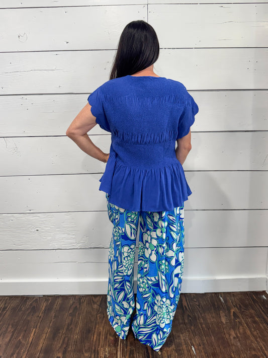 A woman shown from the back wearing a bright blue peplum short-sleeve top and blue floral wide-leg pants, standing against a white wood-paneled wall on a dark wood floor.