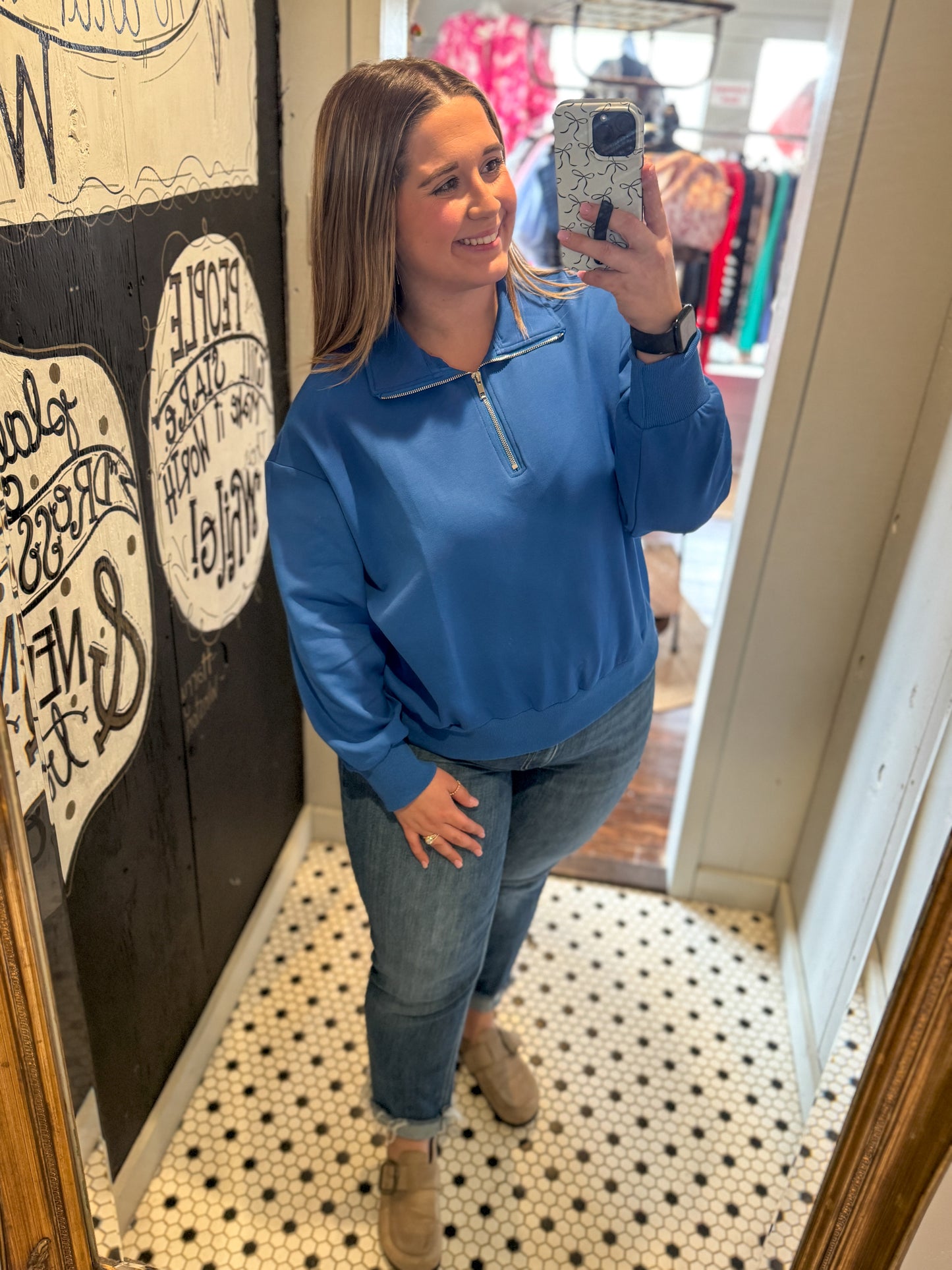 Woman taking a mirror selfie wearing a blue sweatshirt and jeans in a store.