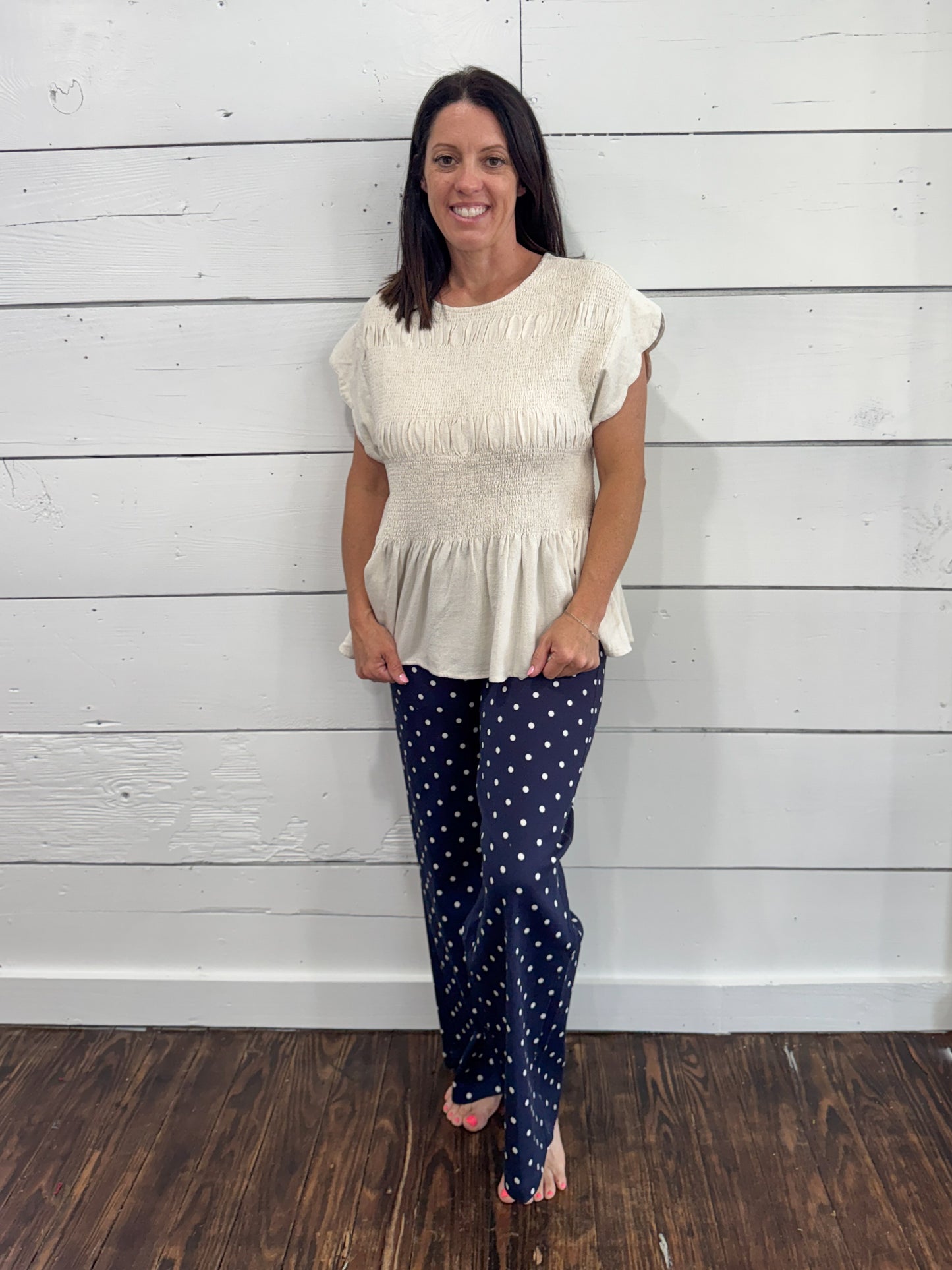 A woman modeling a cream-colored short-sleeve peplum top and navy blue pants with white polka dots, standing against a white wood-paneled wall on a dark wood floor.