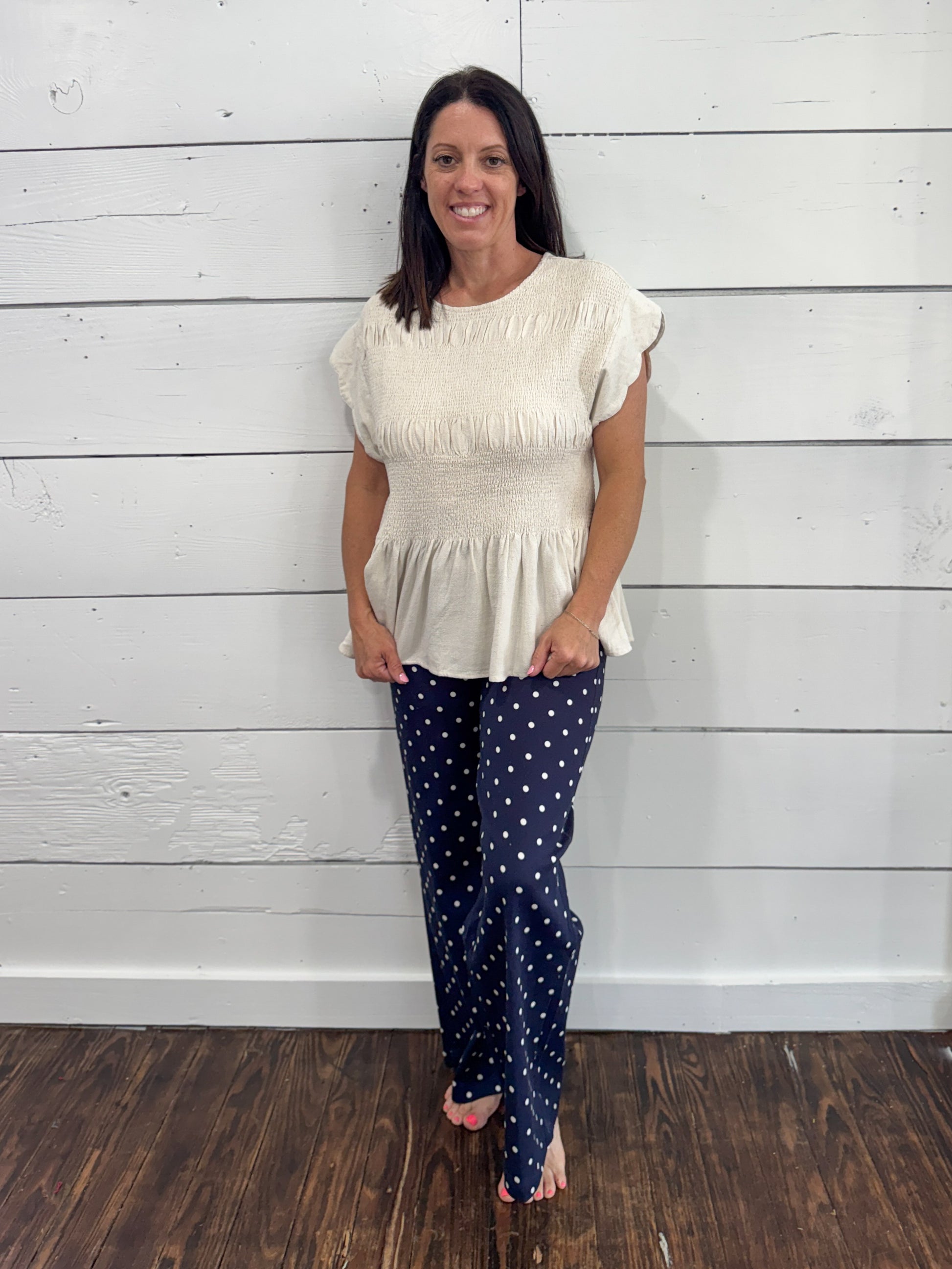 A woman modeling a cream-colored short-sleeve peplum top and navy blue pants with white polka dots, standing against a white wood-paneled wall on a dark wood floor.