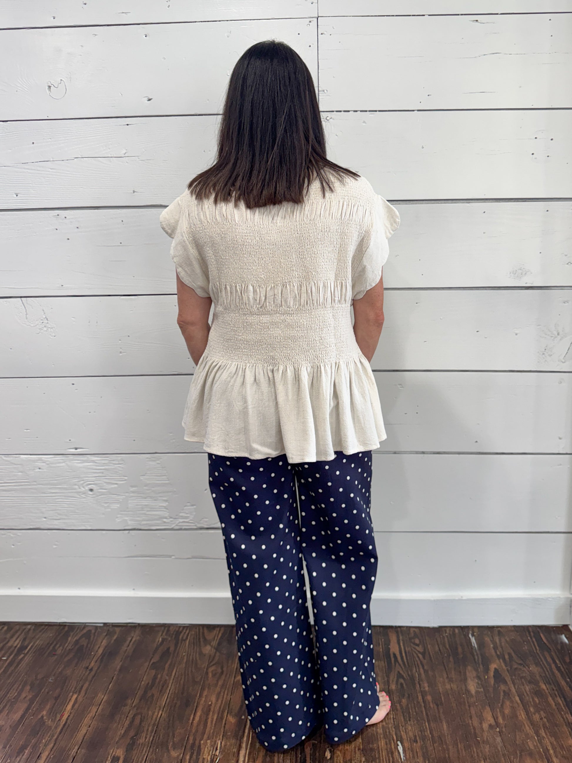 A woman viewed from behind wearing a cream-colored textured peplum top and navy blue wide-leg pants with white polka dots, standing against a white wood-paneled wall on a dark wood floor.