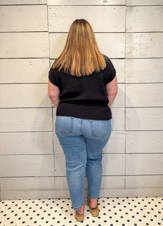 Person wearing a black top and blue jeans standing against a light wooden paneled wall.