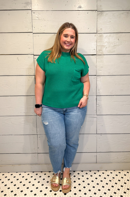 Woman wearing a green top and blue jeans standing against a light wooden paneled wall.