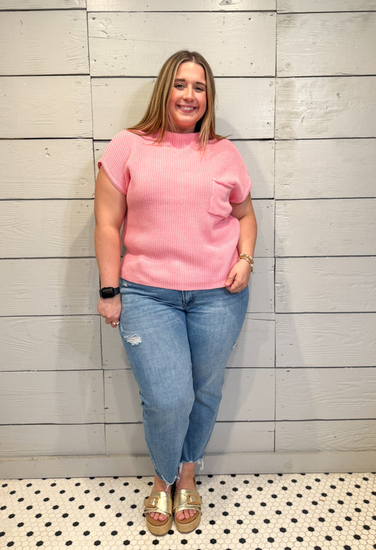 Woman wearing a pink sweater and blue jeans standing against a light wooden paneled wall.